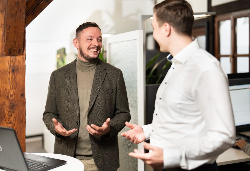 Zwei Männer führen ein freundliches Gespräch in einem modernen Büro, gestikulieren offen und lächeln sich an.