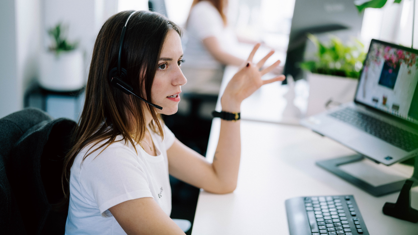 Ihre Webseite in besten Händen Frau mit Headset sitzt am Schreibtisch vor einem Computer und spricht gestikulierend während eines Online-Meetings oder Kundengesprächs.