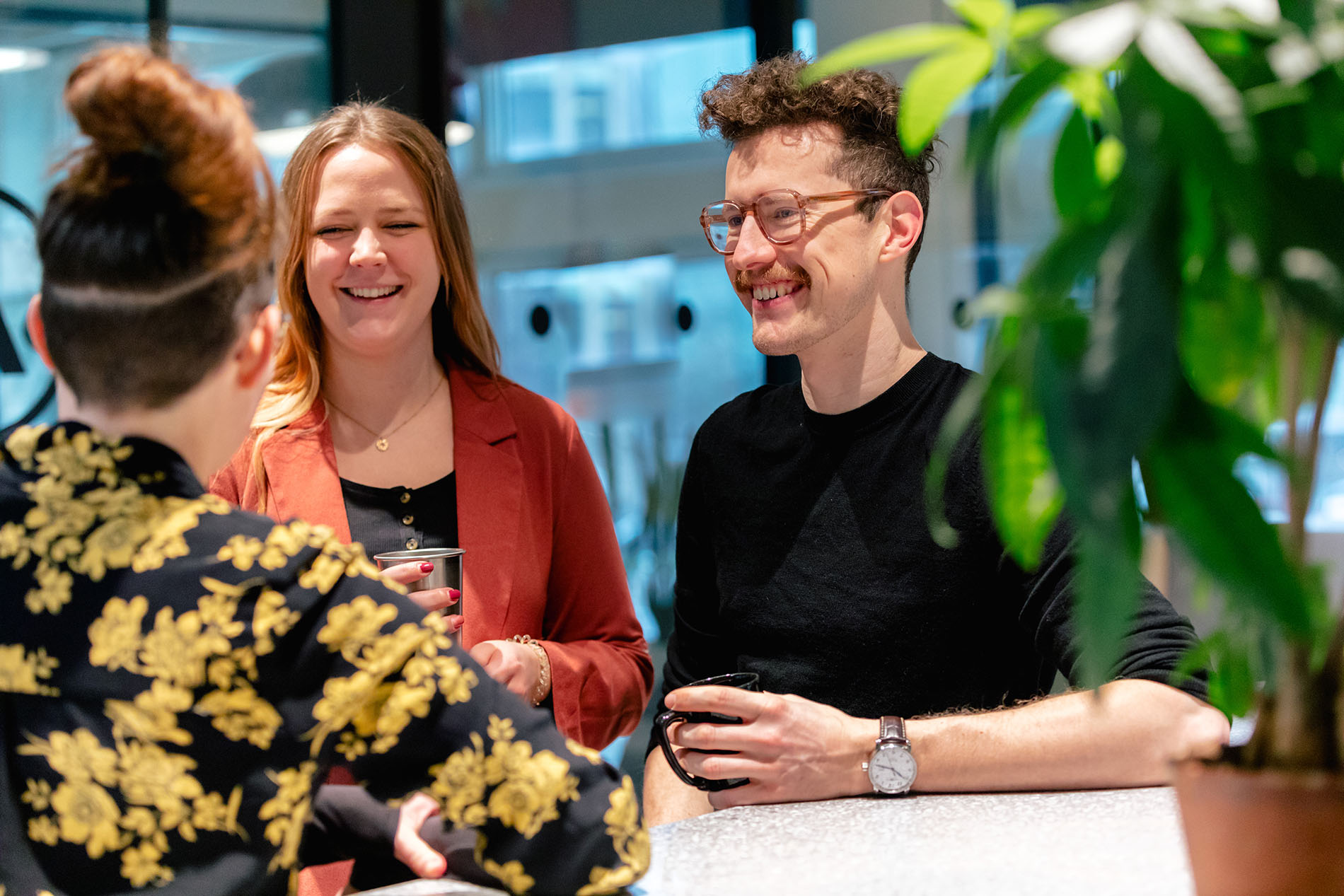 Eine stilvoll gekleidete Gruppe von Menschen unterhält sich, mit Getränken in der Hand und Grünzeug im Hintergrund.